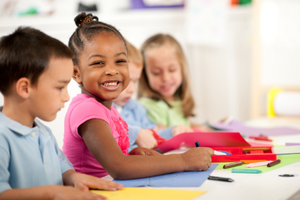 young children smile and sit at a table drawing and writing on construction paper