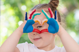a little girl with colorfully painted palms smiles and holds her fingers in the shape of a heart in front of her eye.