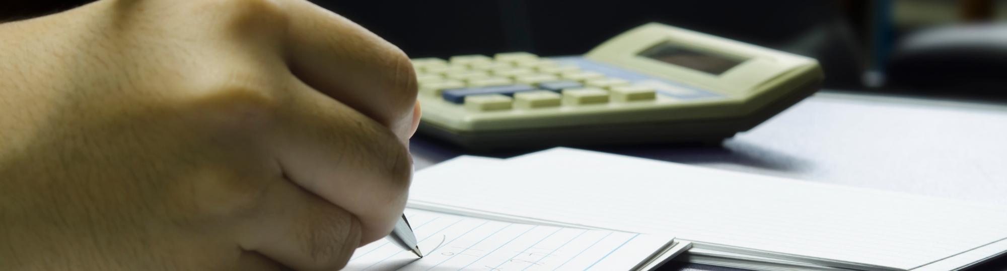 a hand holding a pen doing calculations near a calculator