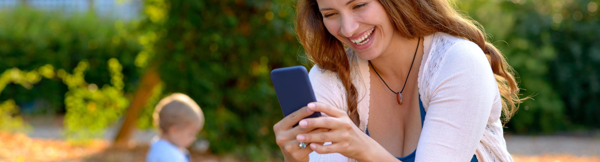 Mother in park smiling at cell phone with baby sitting on ground in background