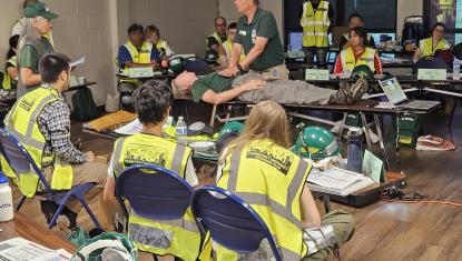 A group of people in CERT training wearing CERT yellow vests watch an instructor demonstrating something with another individual laying on the table.