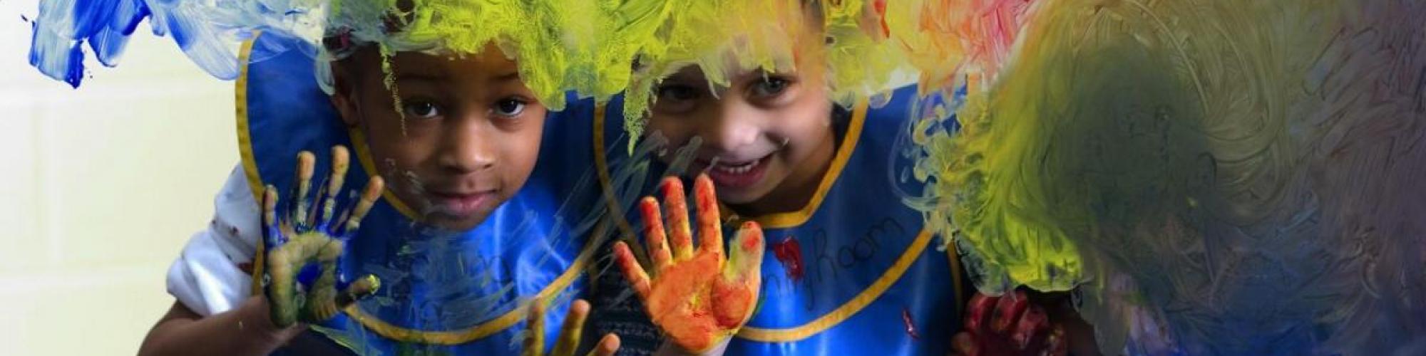 Two children peer through a glass wall where they have been painting with their hands