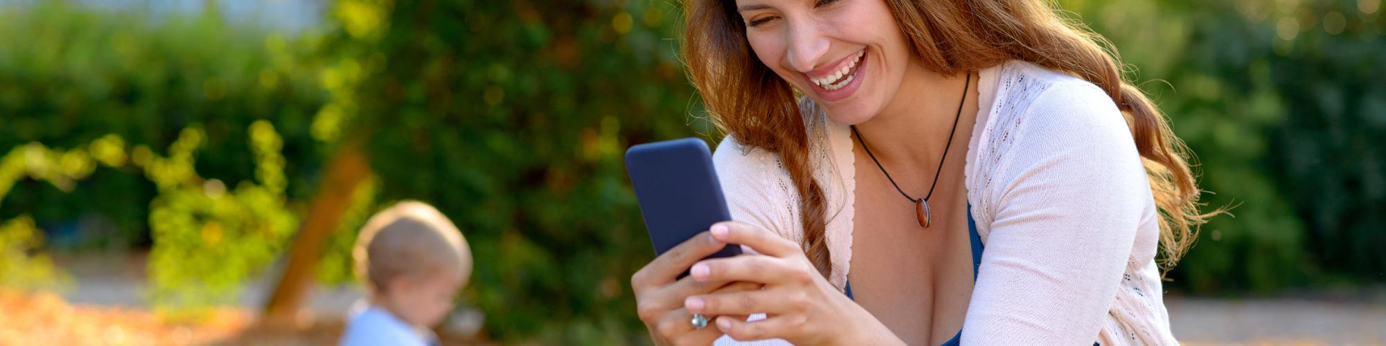 Mother in park smiling at cell phone with baby sitting on ground in background